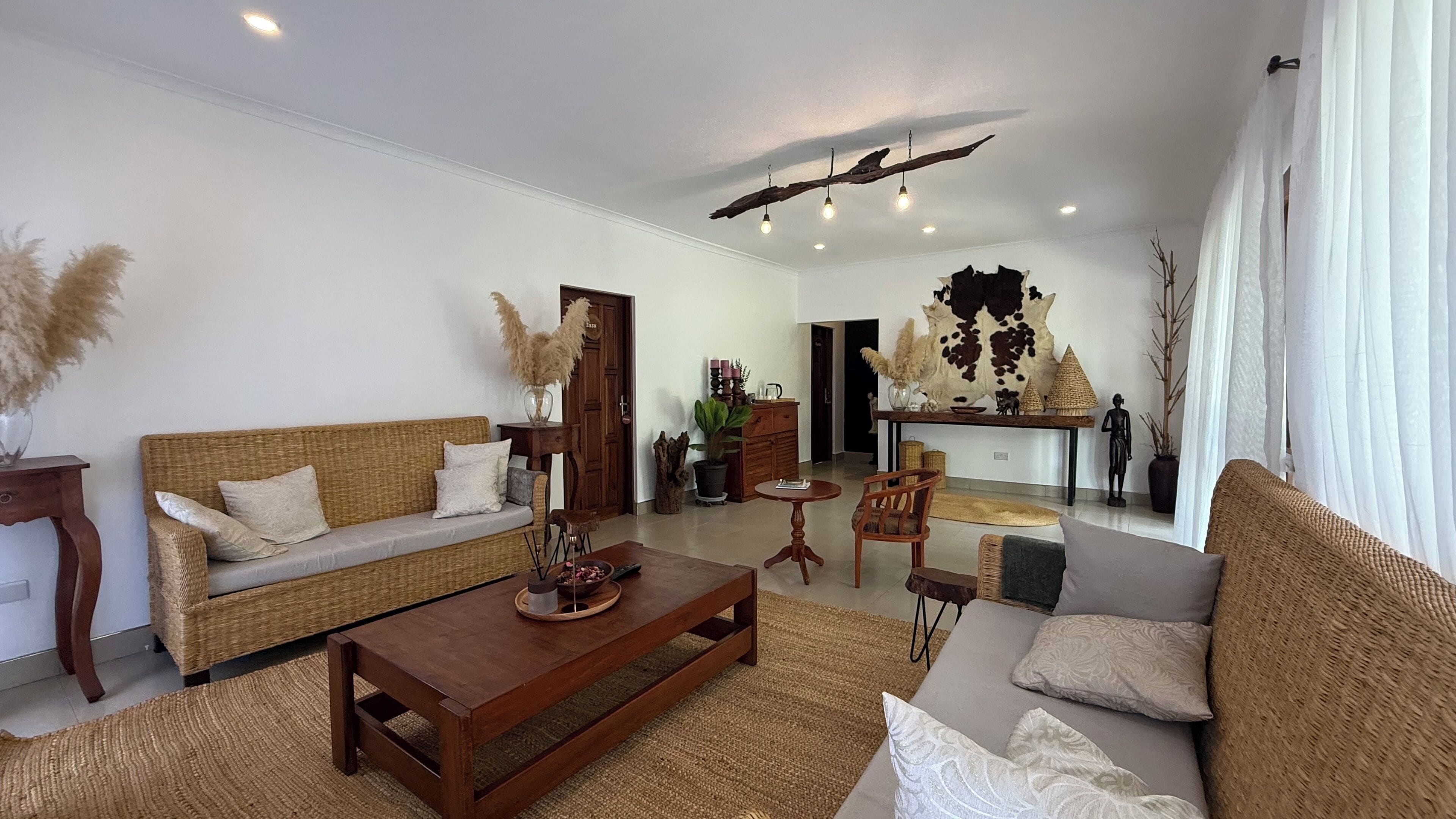 A spacious living room with a sofa, coffee table, and chairs. There is a wooden cabinet and a decorative wall piece.