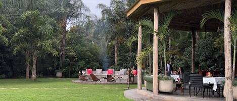 An outdoor seating area with a thatched roof pavilion, surrounded by lush greenery and a well-maintained lawn.