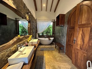 A bathroom with a stone wall, wooden beams, a large mirror, a bathtub, a sink, and a wooden door. - Tulivu Kilimanjaro Retreat (Moshi)