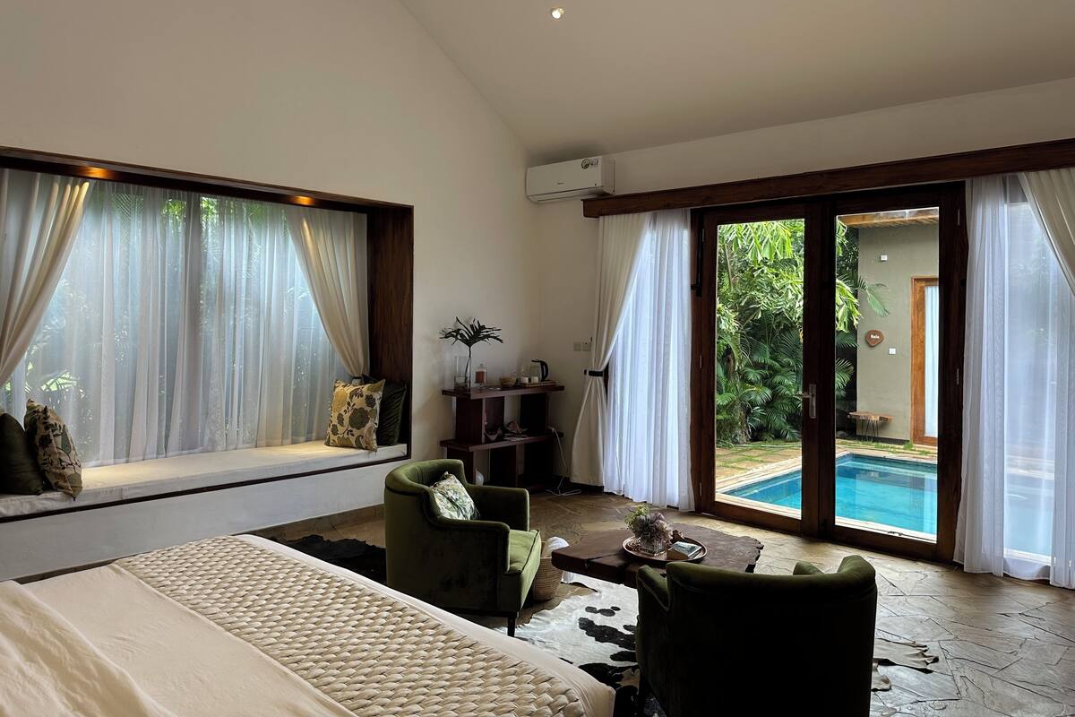 A spacious room with a bed, two armchairs, a coffee table, and a view of a swimming pool.