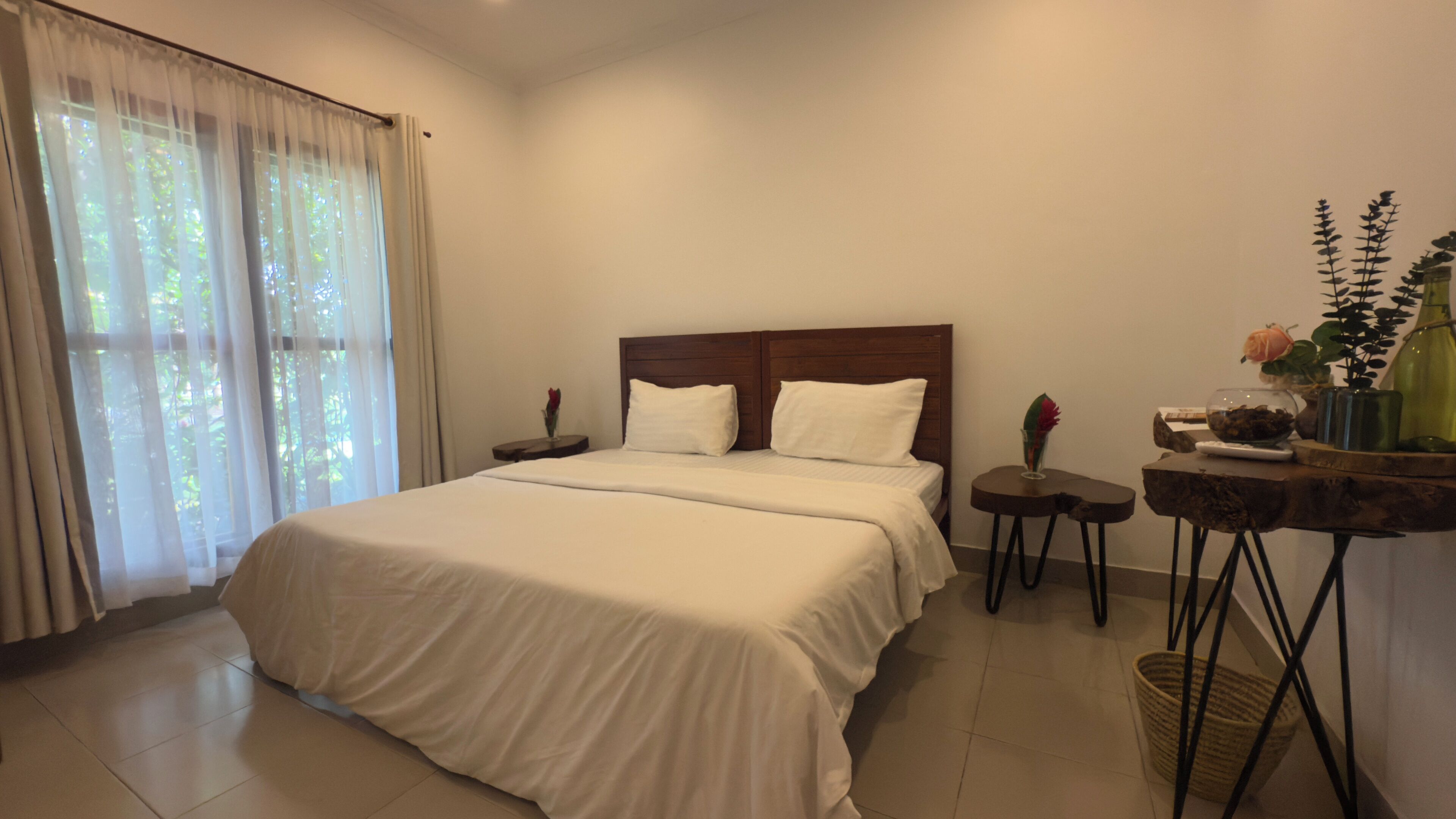 A hotel room with a large bed, a nightstand, a small table with a vase, and a window with curtains.