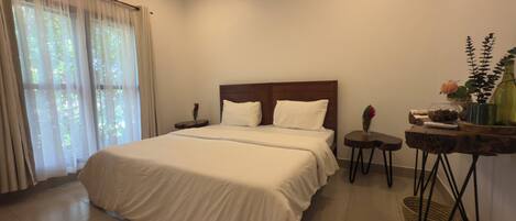 A hotel room with a large bed, a nightstand, a small table with a vase, and a window with curtains.