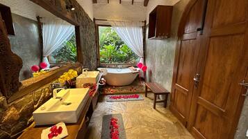 Un baño con bañera, un espejo grande, un tocador de madera y piso de piedra.