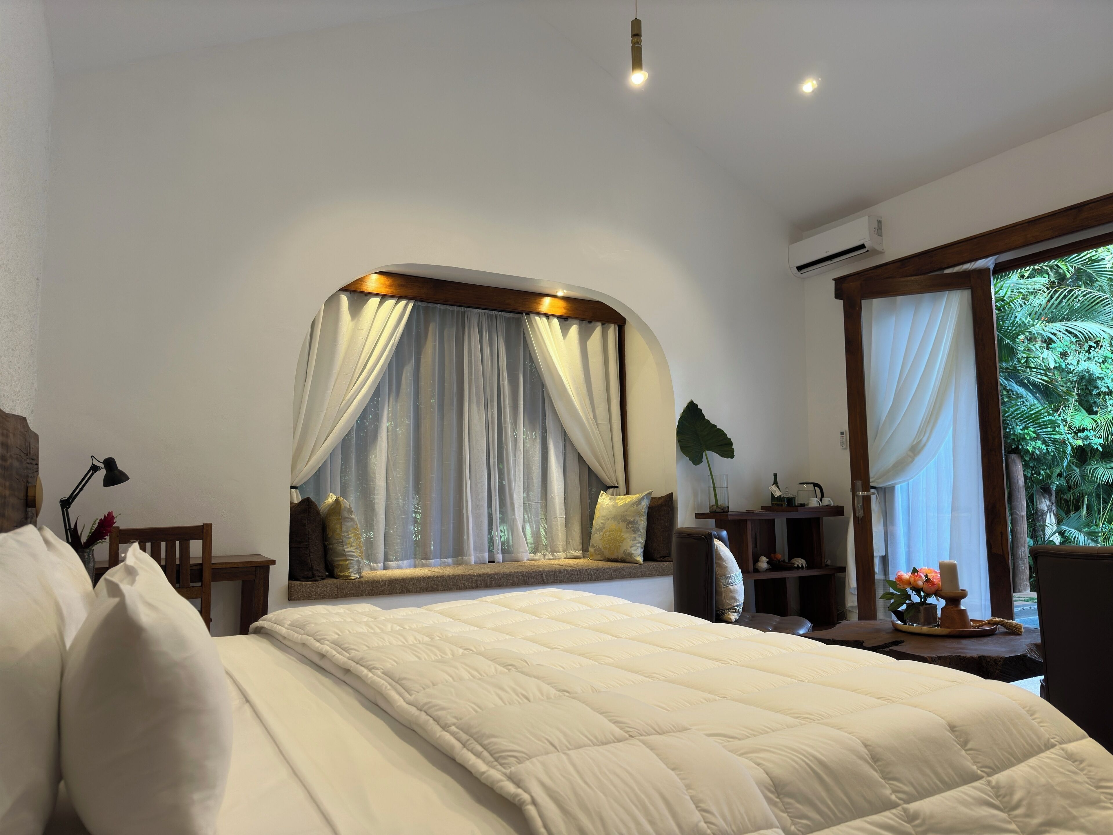 A bedroom with a large bed, a window with curtains, a bedside table with a lamp, and a view of greenery outside.