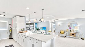Une cuisine moderne avec des meubles blancs, un grand îlot central, des luminaires suspendus et une vue sur l’espace de vie.