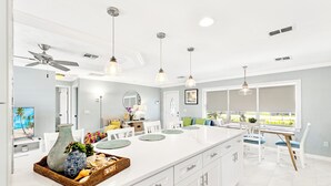 Une cuisine moderne avec des meubles blancs, un grand îlot central, des luminaires suspendus et une vue sur l’extérieur.