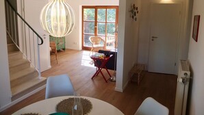 Een ruime woonkamer met een trap, een eettafel met een geweven placemat en een groot raam met uitzicht op groen.