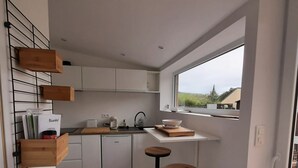 A modern kitchen with white cabinets, a built-in oven, a coffee machine, and a window overlooking a green landscape. - Très Joli Studio, Neuf, à 50 m de la mer (Paimpol)