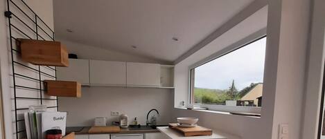 A modern kitchen with white cabinets, a built-in oven, a coffee machine, and a window overlooking a green landscape.