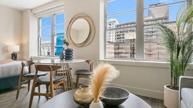 Une chambre d’hôtel moderne avec une grande fenêtre, un miroir rond, une table à manger avec des chaises, un lit et une plante en pot.