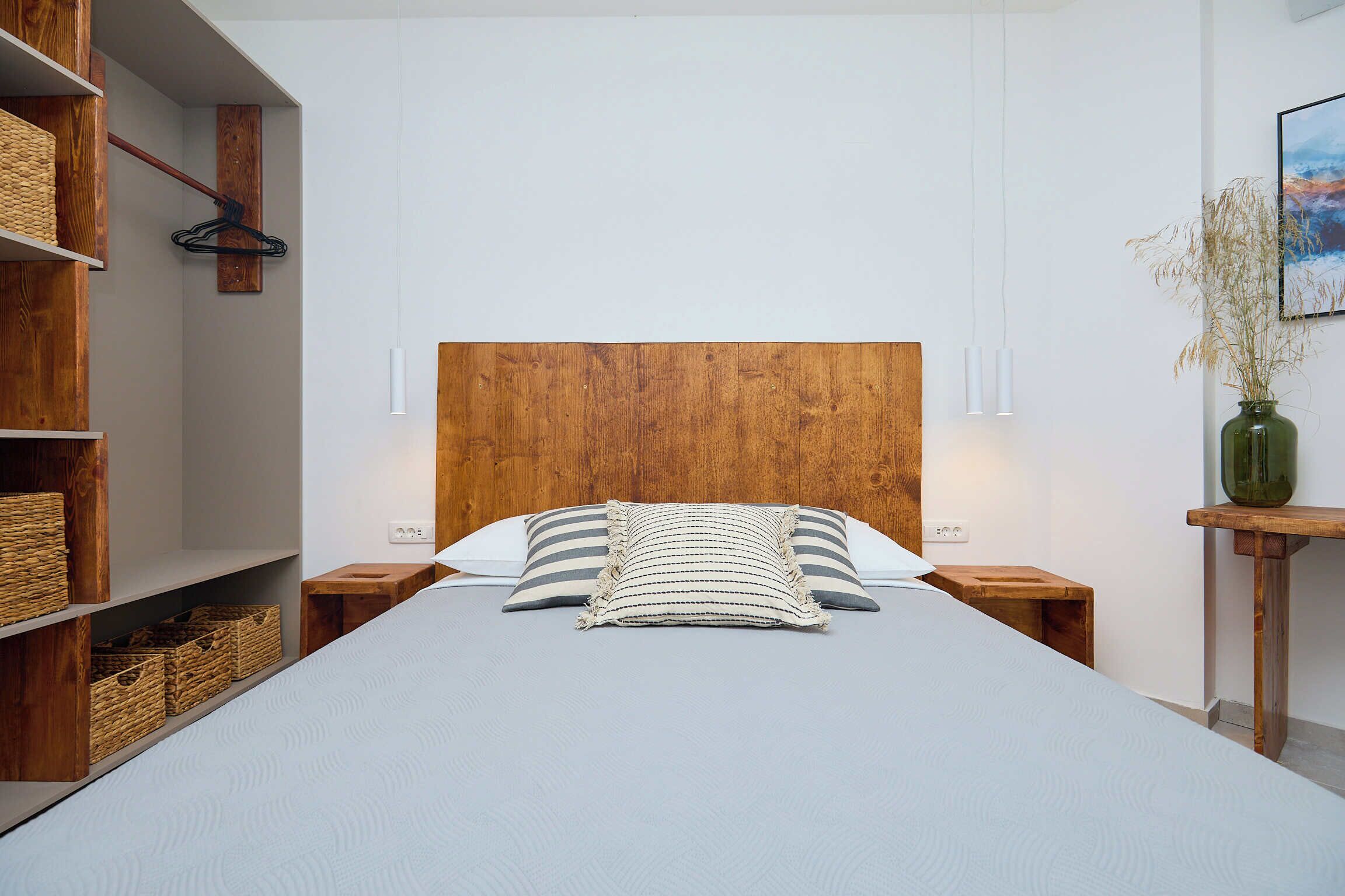Una cama bien hecha con cabecera de madera, dos mesitas de noche y un pequeño taburete de madera.