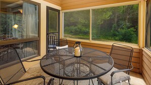 Un patio cubierto con una mesa redonda de metal y cuatro sillas, una jarrilla sobre la mesa, y amplios ventanales que dan a un bosque.