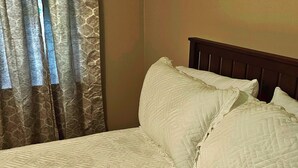 Un lit recouvert d’une courtepointe blanche matelassée, avec deux oreillers, une tête de lit en bois et une fenêtre ornée de rideaux à motifs.
