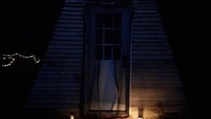 Une petite cabane en bois sombre, avec une seule fenêtre et une porte, entourée d’arbres.
