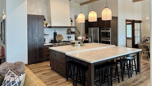 Une cuisine moderne dotée d’un grand îlot central, de meubles de rangement en bois foncé, d’appareils électroménagers en acier inoxydable et de luminaires suspendus.
