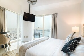 A hotel room with a large bed, a TV mounted on the wall, a desk with a chair, and a view of a cityscape through large windows.