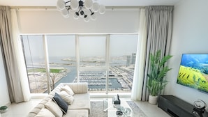 A modern living room with a large window, a sofa, a coffee table, a TV, and a plant. - OSKENA Homes-Damac Heights Two Bedroom (Dubai)