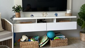 Un salon doté d’un téléviseur à écran plat fixé sur un meuble de rangement blanc, entouré d’étagères où sont disposées des plantes en pot, d’un panier en osier et d’un panier tressé contenant du matériel de sport.