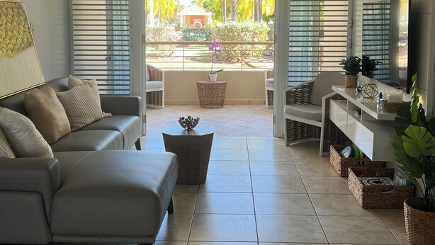 Un salon moderne avec un canapé gris, un ventilateur de plafond et une vue sur un jardin grâce à de grandes fenêtres ouvertes.