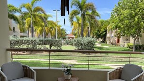 Un balcon avec des fauteuils en osier, une petite table agrémentée d’une plante, et une vue sur un jardin bien entretenu, avec des palmiers et un bâtiment en arrière-plan.