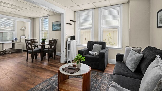 A spacious living room with a dark grey sofa, a black armchair, and a wooden coffee table. There is a dining area with a table and chairs, and large windows with blinds. - 2BR Near Duluth + Spacious & Work‑Friendly (Superior)