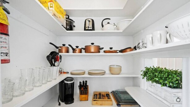 Une cuisine avec des étagères blanches, une machine à café, des casseroles, un grille-pain et une plante.