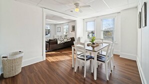 Une pièce spacieuse comprenant une table à manger, des chaises, un ventilateur de plafond et donnant sur un salon avec un canapé et des fenêtres.