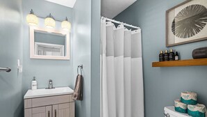 Une salle de bain avec un lavabo blanc, un miroir et un rideau de douche. On trouve des articles de toilette sur le lavabo et un porte-rouleau de papier toilette au mur.