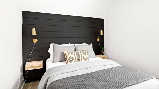Un dormitorio moderno con cabecera negra, dos mesitas de noche, una cama con ropa de cama gris y dos lámparas fijadas a la pared.