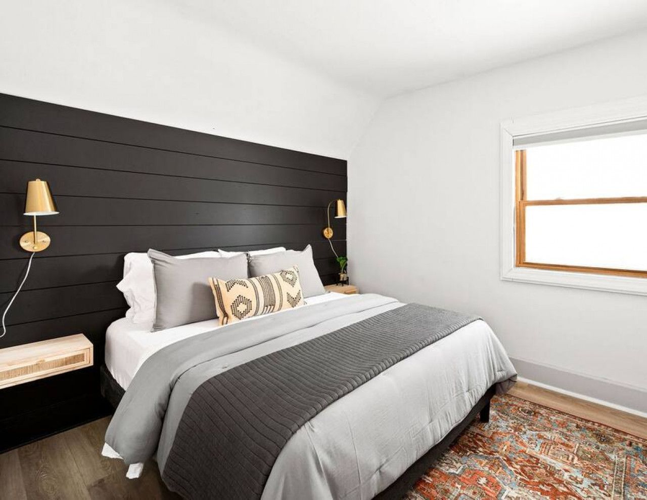 A modern bedroom with a black headboard, a bed with grey bedding, two bedside tables with lamps, and a patterned rug.
