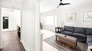 Un salón amplio con un sofá gris, un banco de madera y un ventilador de techo. Hay dos cuadros enmarcados en la pared y un ventanal que deja entrar luz natural.
