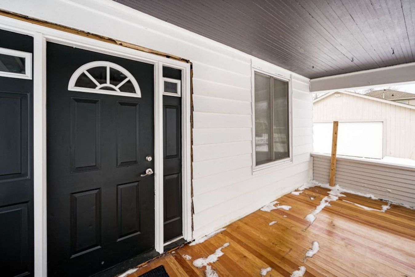 Una casa amb porta principal negra, revestiment blanc i un sòl de fusta amb una mica de neu.