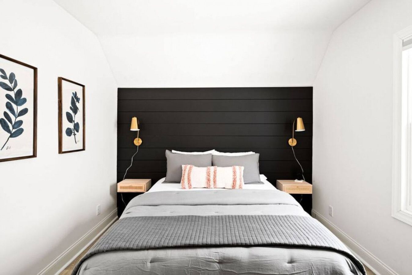 Une chambre à coucher avec une tête de lit noire, un lit avec une literie grise, deux tables de chevet avec des lampes et, au mur, des reproductions encadrées de motifs botaniques.