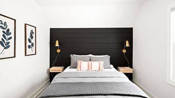 Ein Schlafzimmer mit einem schwarzen Kopfteil, einem Bett mit grauer Bettwäsche, zwei Nachttischen mit Lampen und gerahmten botanischen Drucken an der Wand.