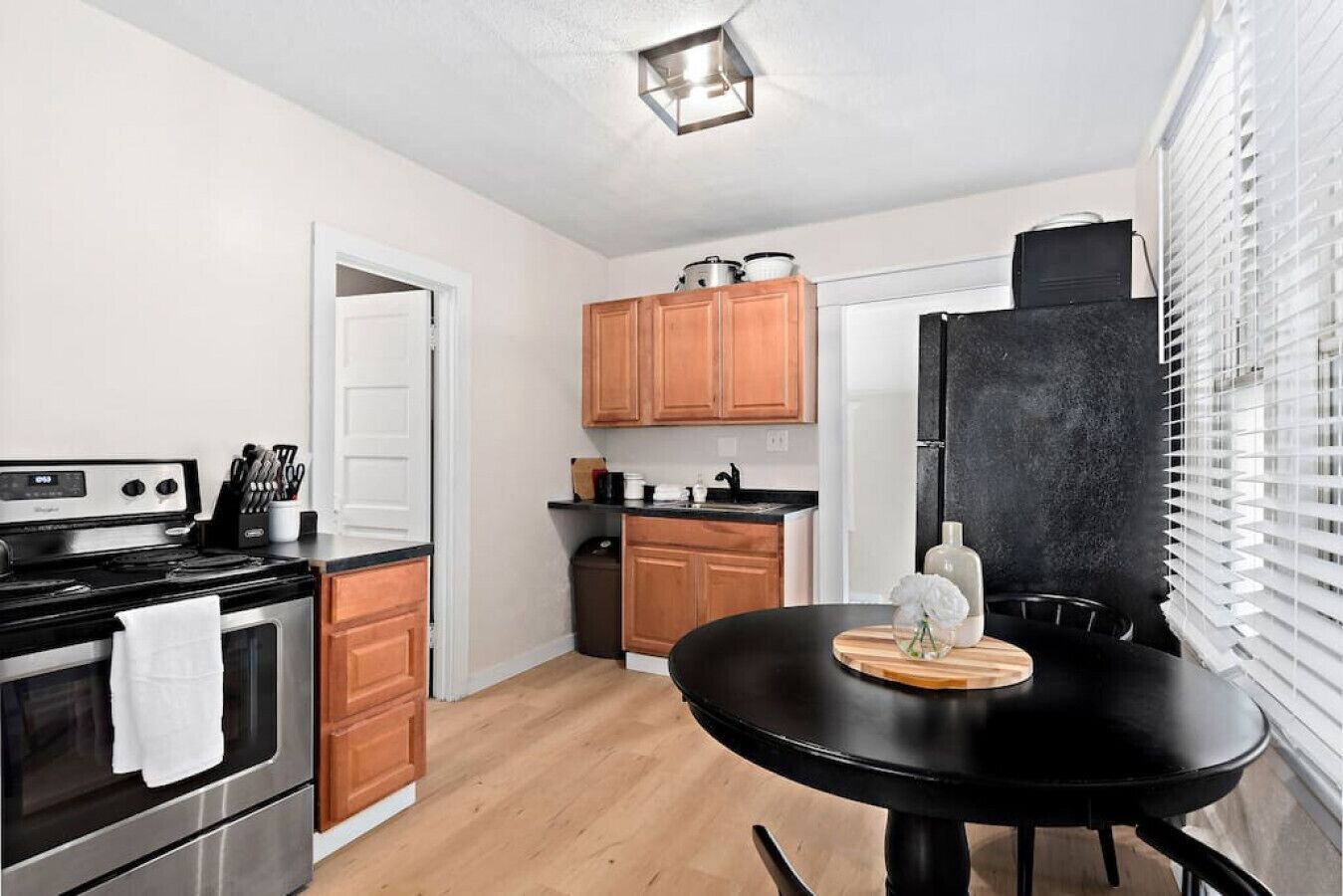 Una cocina compacta con estufa, horno, armarios de madera, un refrigerador negro y una mesa de comedor con un florero.