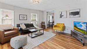 En moderne stue med sofa, lænestol, sofabord og fjernsyn. Der er vinduer med persienner, et spiseområde i baggrunden og kunst på væggene.