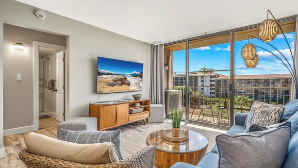 Una sala de estar moderna con un televisor grande, asientos cómodos y un balcón con vistas a edificios y vegetación.
