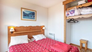 A bedroom with a wooden headboard, two beds with red floral bedding, a framed picture on the wall, and a shelf with folded towels and a package. - Appartement 6 pers, vue montagne avec terrasse (Chamrousse)