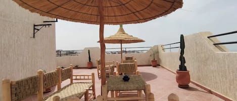 A rooftop terrace with wicker furniture, a large thatched umbrella, and potted plants.