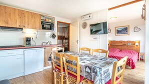 A compact kitchen with a dining area, a TV, and a bed with a red bedspread.