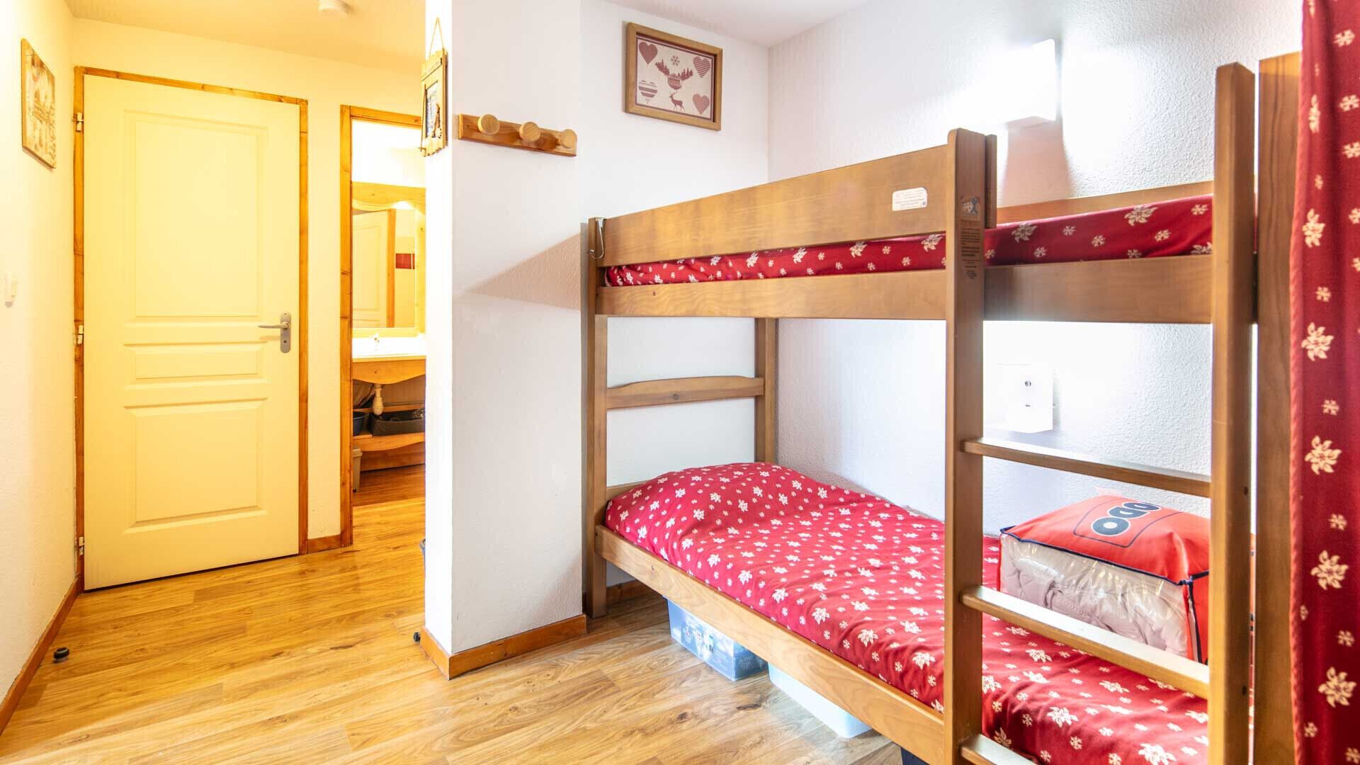 Een slaapkamer met een stapelbed met houten frame, een rode bloemenprint sprei, en een deur naar een andere kamer.