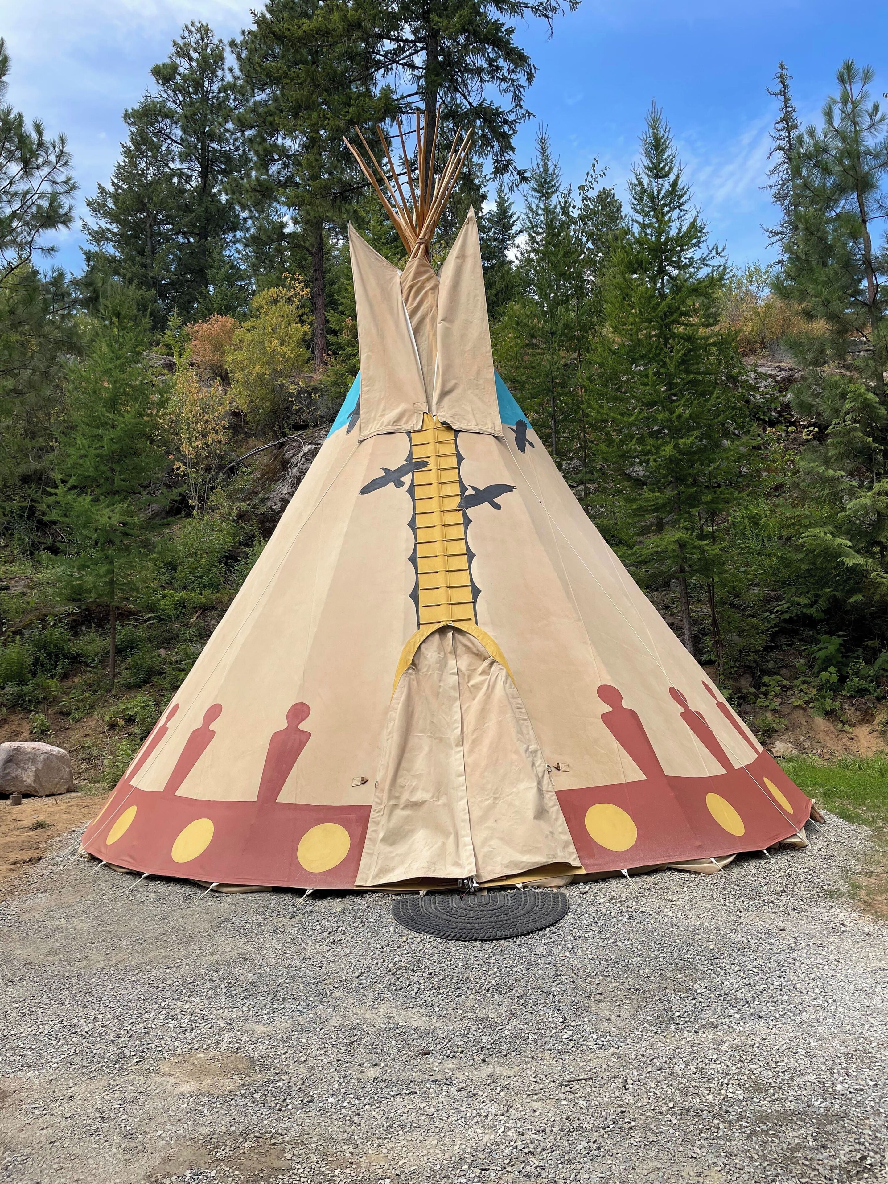 Une grande tente conique beige, ornée de motifs rouges et jaunes, est installée sur un sol de gravier, avec des arbres en arrière-plan.