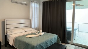 A hotel room with a bed, a ceiling fan, an air conditioner, a window with curtains, and a view of the outside.