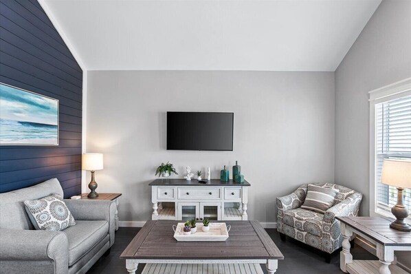 A living room with a grey sofa, patterned armchair, and a coffee table with decorative items. A white console table with plants and decorative objects is against the wall.