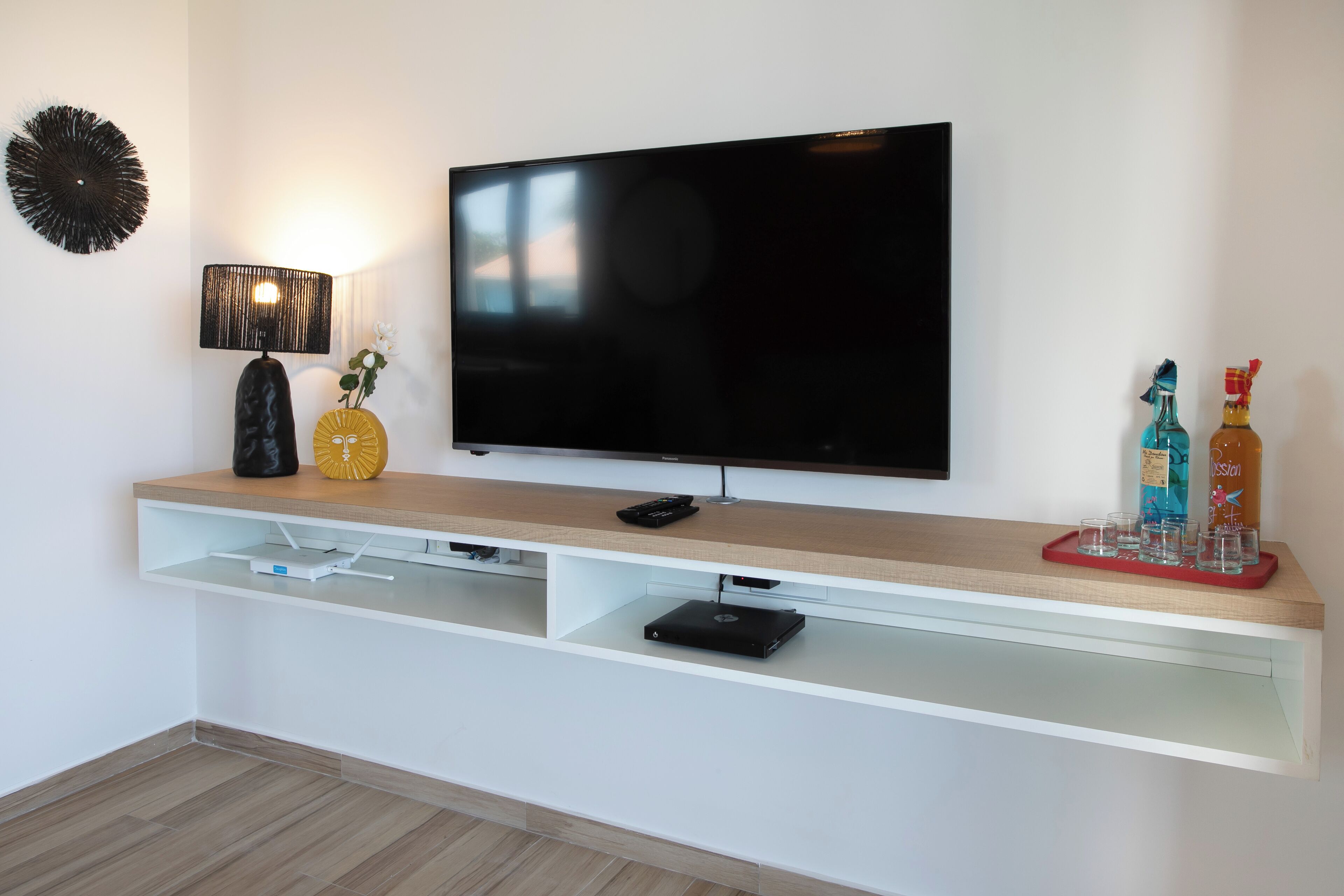 A modern living room with a flat-screen TV mounted on a white wall, a wooden console table with a remote control, a decorative lamp, and a tray with glasses and bottles.