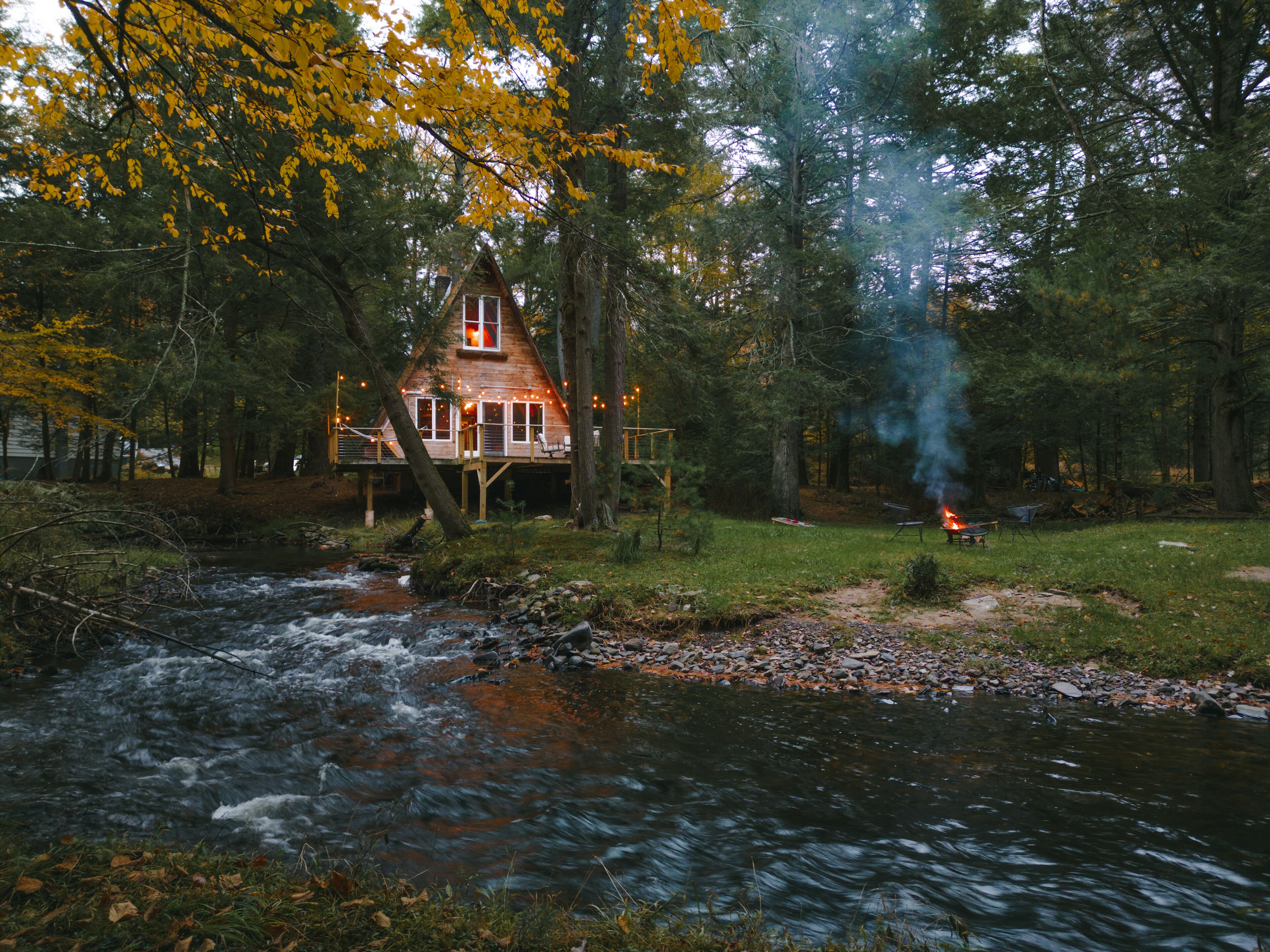 Een blokhut met terras, uitzicht op een rivier, omringd door bomen en een kampvuur.