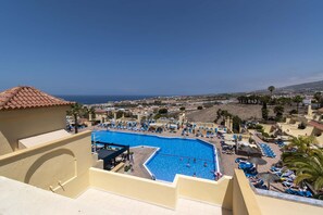 A resort with a large swimming pool surrounded by sun loungers and umbrellas, a clear view of the sea, and a hilly landscape in the background. - Horizon 1 (Costa Adeje)