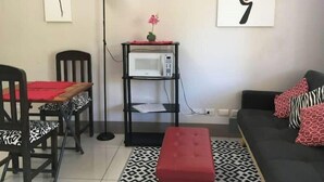 A compact living space with a dining area, a sofa, a red ottoman, a standing lamp, and a microwave on a black shelf.