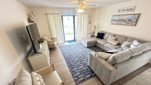 A modern living room with a grey sectional sofa, a glass desk, a wicker chair, and a patterned rug. - Ground floor unit! Close to pool and beach. (Saint George Island)