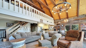 A spacious living room with a high ceiling, wooden beams, and a balcony. There are two sofas, a coffee table, and a patterned armchair. - Tranquil Hillside Home w/ Private Hot Tub! (Pioneer)